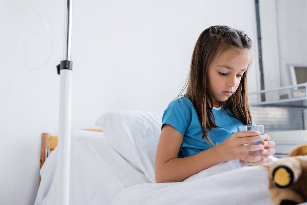 Ill kid holding glass of water near blurred toy on bed in clinic  - 写真・画像