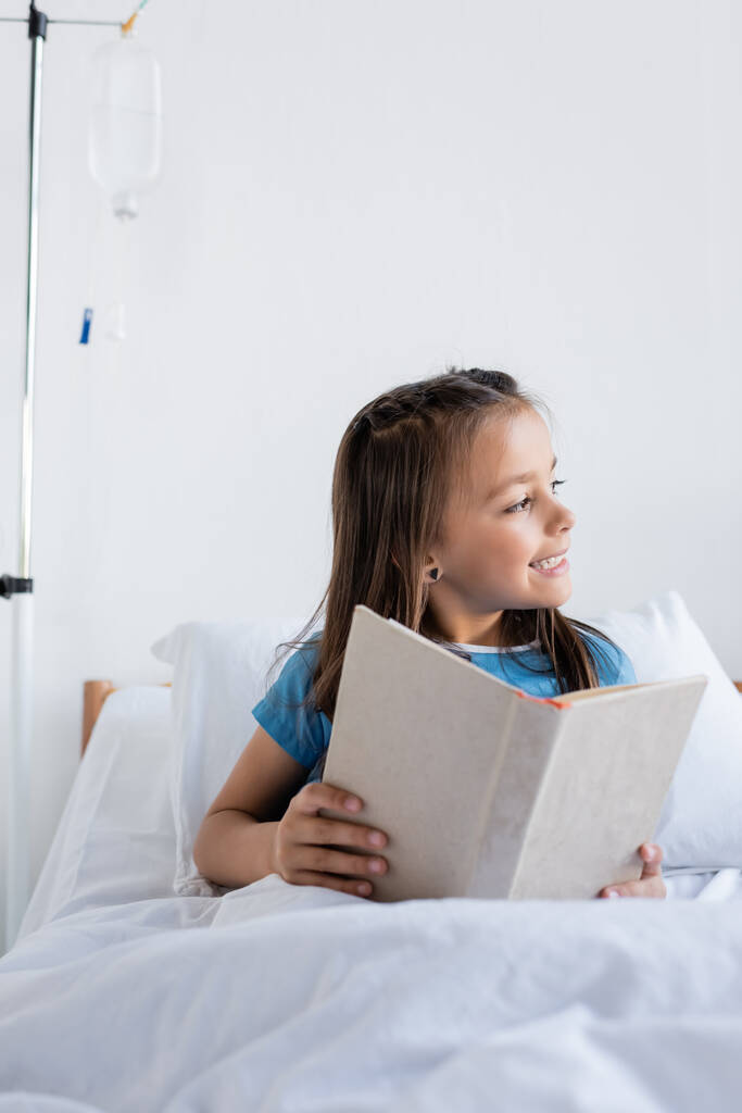 Glimlachend kind met boek op bed op ziekenhuisafdeling  - Foto, afbeelding
