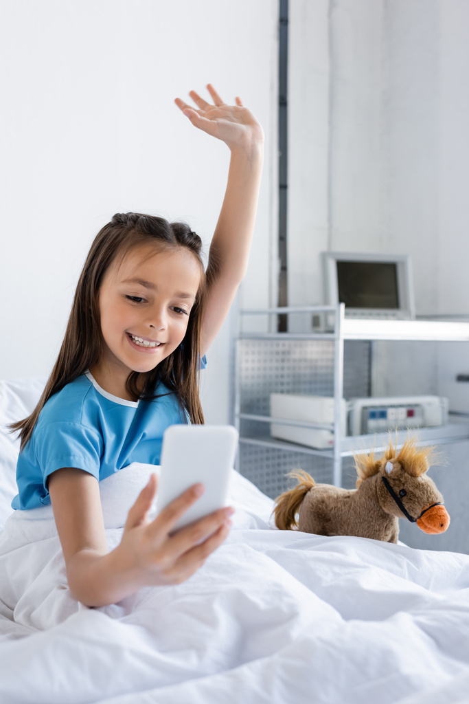 Θετικό παιδί σε φόρεμα ασθενή έχει βιντεοκλήση στο smartphone κοντά σε μαλακό παιχνίδι στο κρεβάτι στην κλινική  - Φωτογραφία, εικόνα