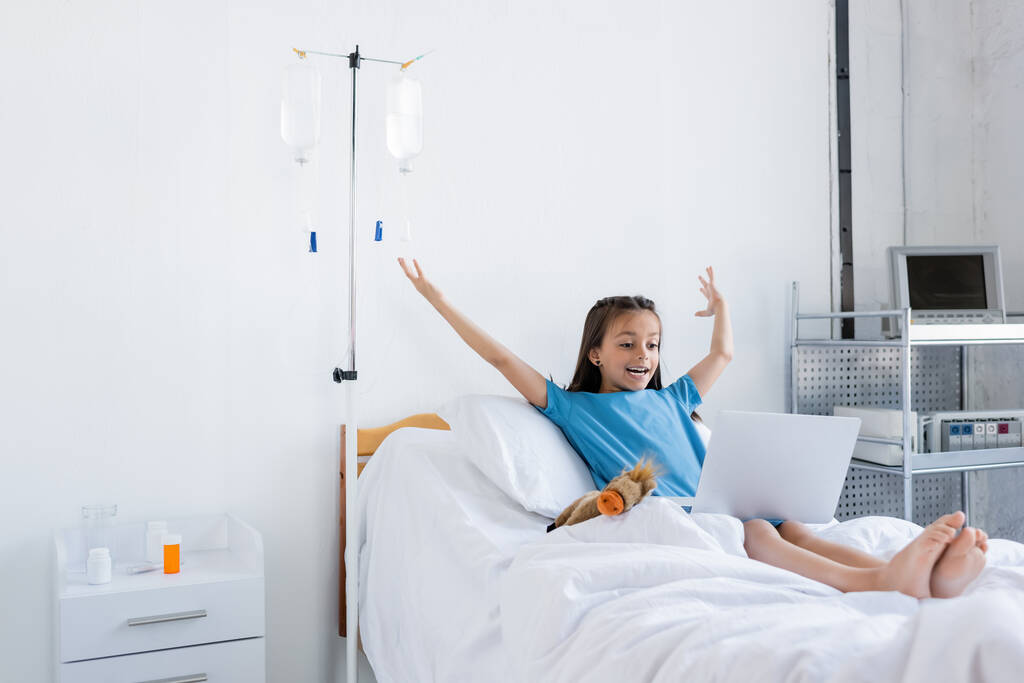 Excited kid using laptop near toy on bed in clinic  - Photo, Image