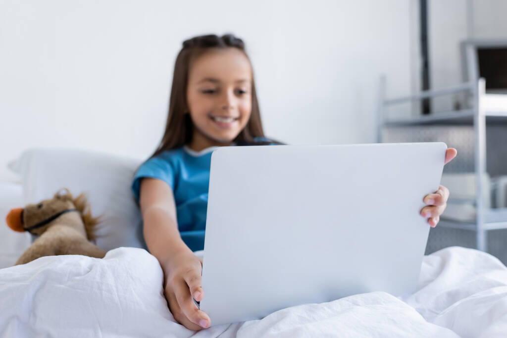Homályos gyerek betegköpenyben laptoppal közel játék az ágyon a kórházban  - Fotó, kép