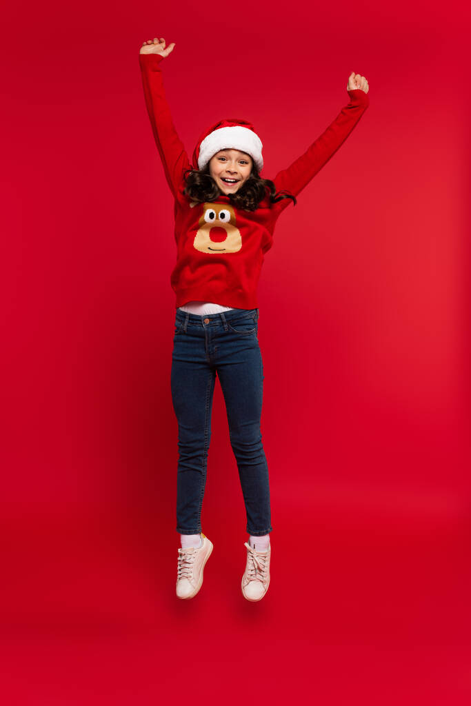 full length of happy girl in santa hat and sweater levitating on red  - Photo, Image