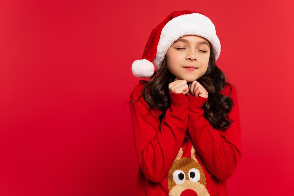 happy girl with closed eyes standing in santa hat and Christmas sweater isolated on red - Photo, Image