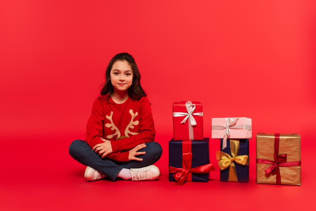 full length of cheerful kid in winter sweater and jeans sitting near Christmas presents on red - Photo, Image