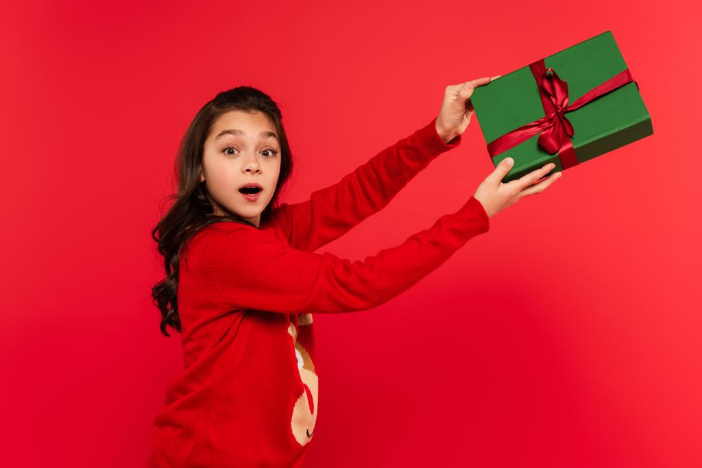 choqué fille en hiver pull tenant enveloppé cadeau de Noël isolé sur rouge - Photo, image
