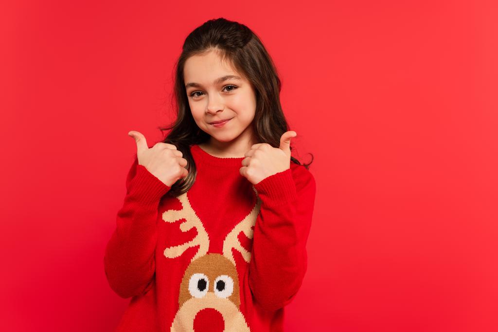 positief meisje in de winter trui tonen duimen omhoog en kijken naar camera geïsoleerd op rood - Foto, afbeelding