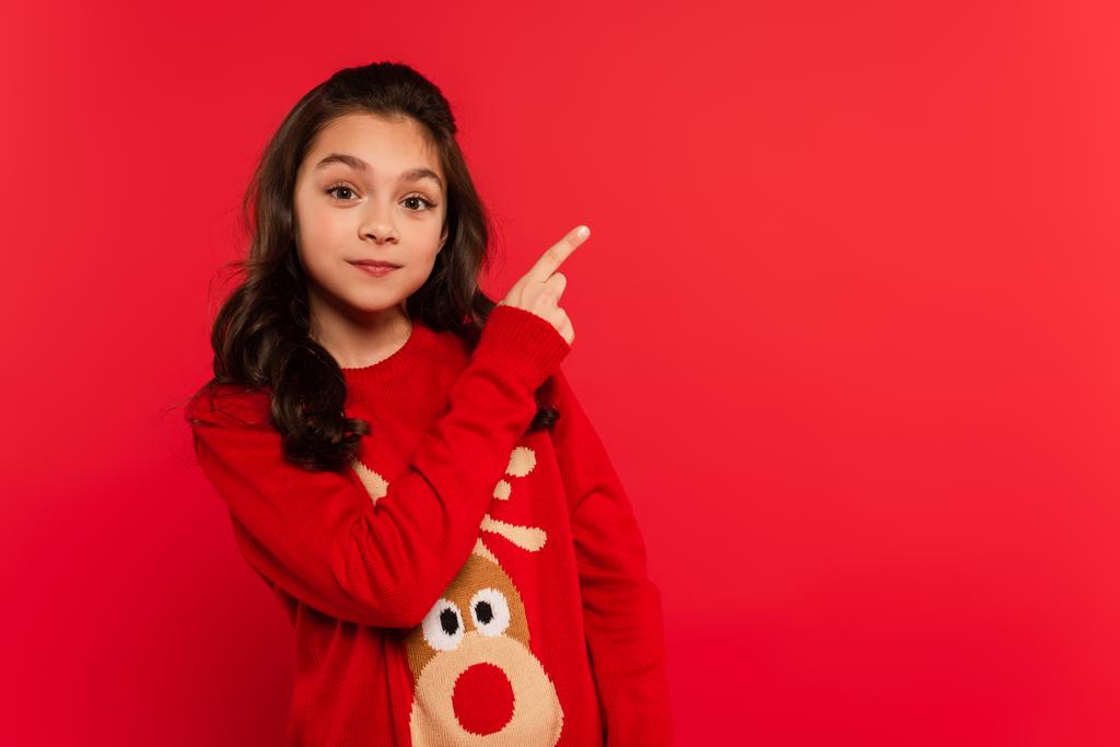 χαρούμενο παιδί με χριστουγεννιάτικο πουλόβερ που δείχνει με το δάχτυλο απομονωμένο στο κόκκινο - Φωτογραφία, εικόνα
