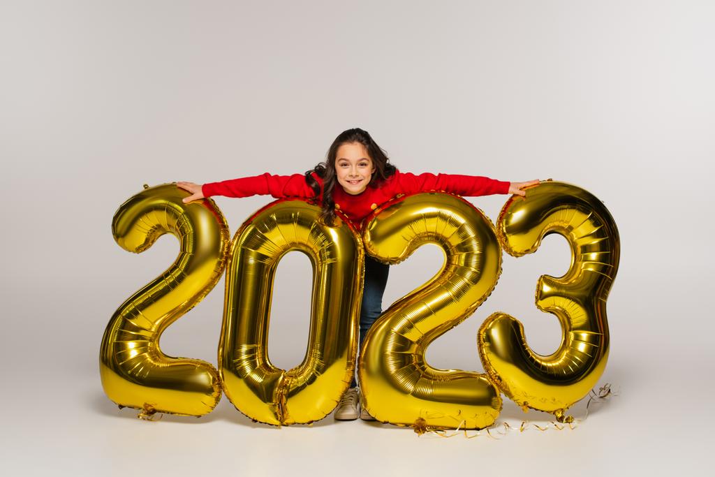 volledige lengte van vrolijk kind in rode trui knuffelen ballonnen met 2023 nummers op grijs - Foto, afbeelding