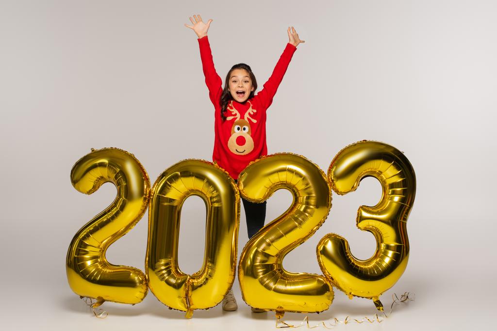 Staunendes Kind in rotem Pullover, das mit erhobenen Händen neben Luftballons mit 2023 Ziffern auf grau steht - Foto, Bild