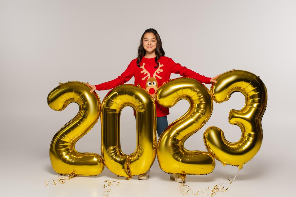 volledige lengte van het lachende meisje in rode trui bij ballonnen met 2023 cijfers op grijs - Foto, afbeelding