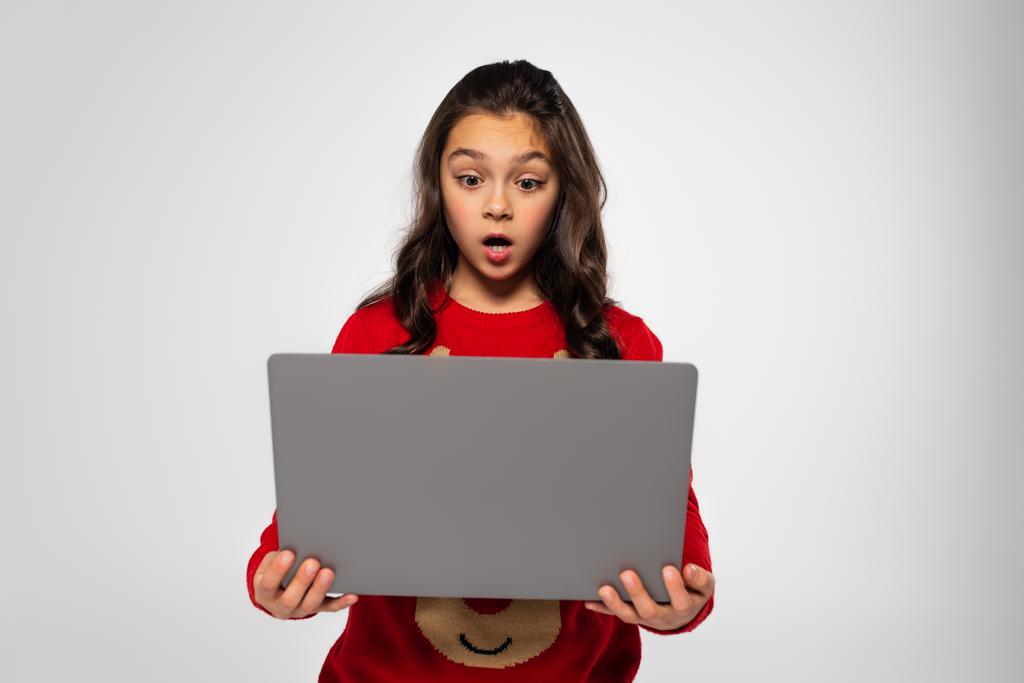 shocked child in red Christmas sweater looking at laptop isolated on grey - Photo, Image