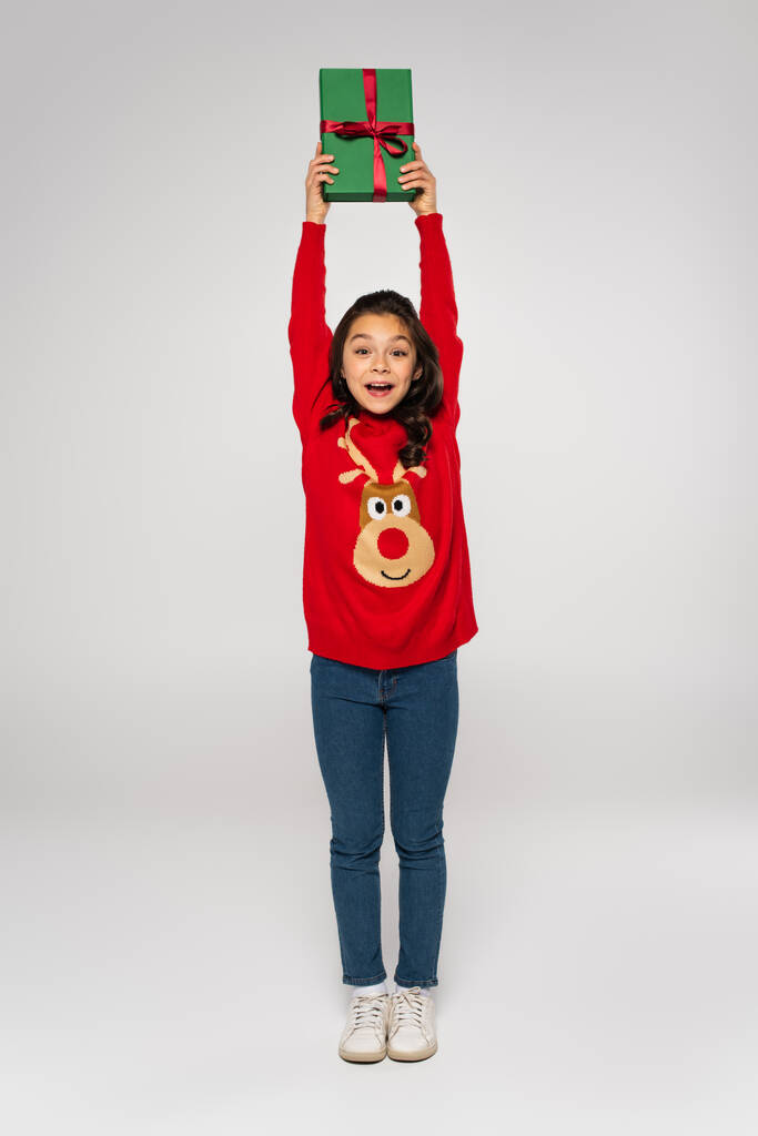 comprimento total da menina animado em camisola vermelha segurando presente de Natal acima da cabeça em cinza - Foto, Imagem