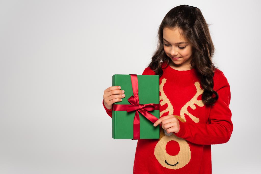 Ευτυχισμένο κορίτσι σε κόκκινο πουλόβερ κοιτάζοντας τυλιγμένο χριστουγεννιάτικο δώρο απομονωμένο σε γκρι - Φωτογραφία, εικόνα