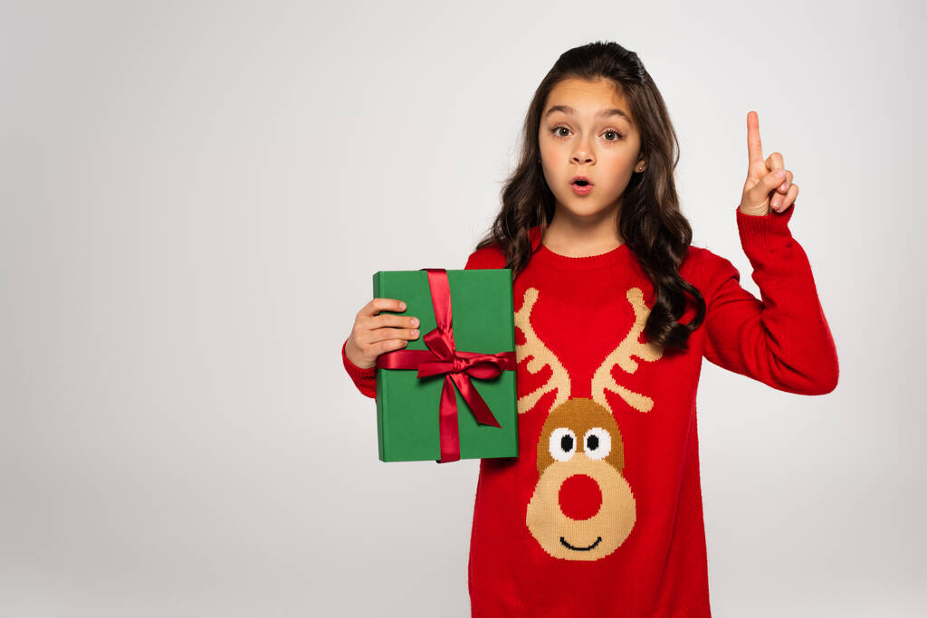 verrast meisje in rode trui houden verpakt kerstcadeau en wijzen met de vinger geïsoleerd op grijs - Foto, afbeelding