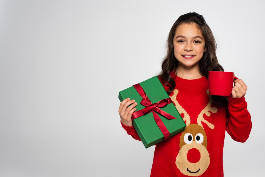happy girl in red sweater holding cup of cocoa and Christmas present isolated on grey - Photo, Image