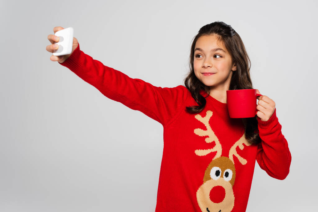 Glimlachend meisje in Nieuwjaar trui holding cup en het nemen van selfie geïsoleerd op grijs  - Foto, afbeelding
