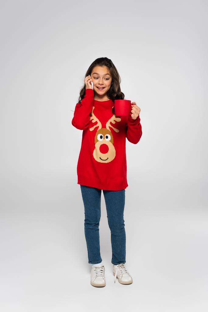Полная длина ребенка в новогоднем свитере, говорящего по смартфону и держащего чашку на сером  - Фото, изображение