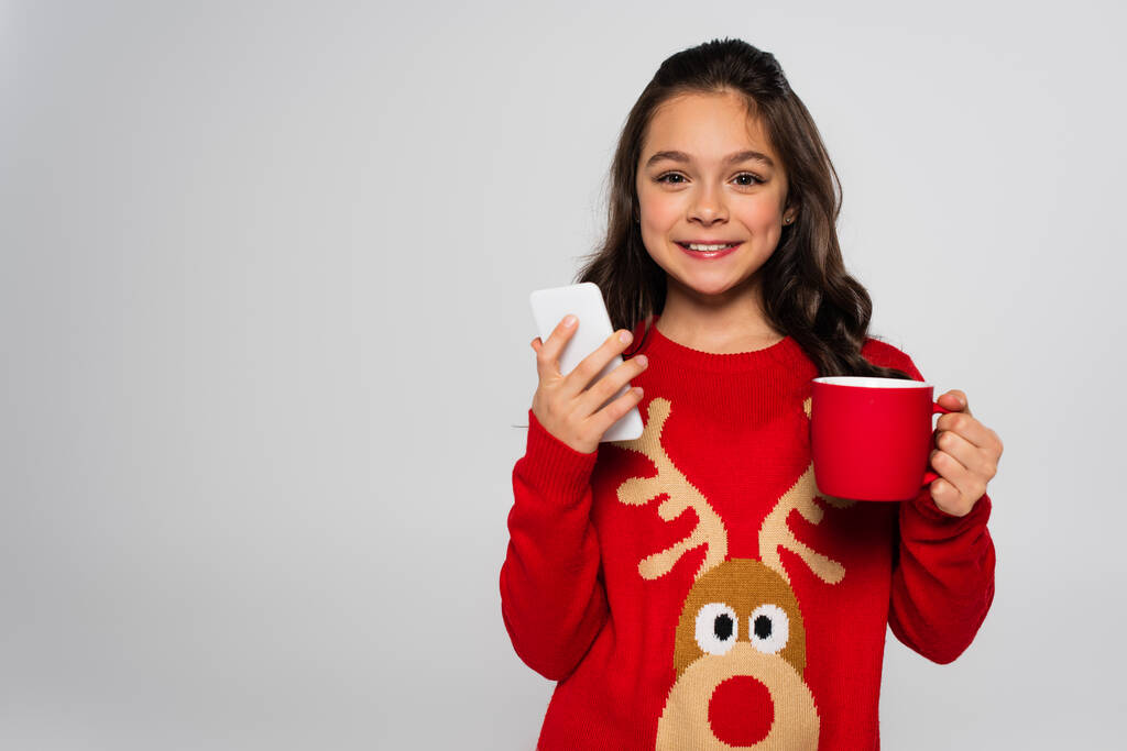 Positive child in New Year sweater holding cup and smartphone isolated on grey  - Photo, Image