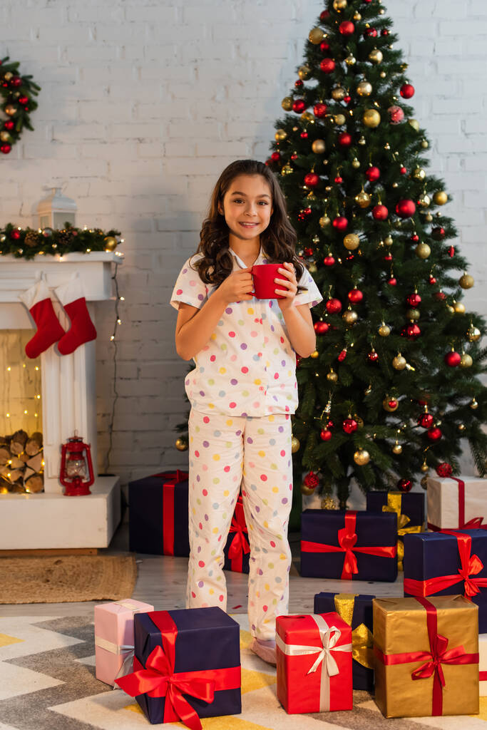 Uśmiechnięty dzieciak w piżamie trzyma kubek w pobliżu choinki i prezenty w domu  - Zdjęcie, obraz
