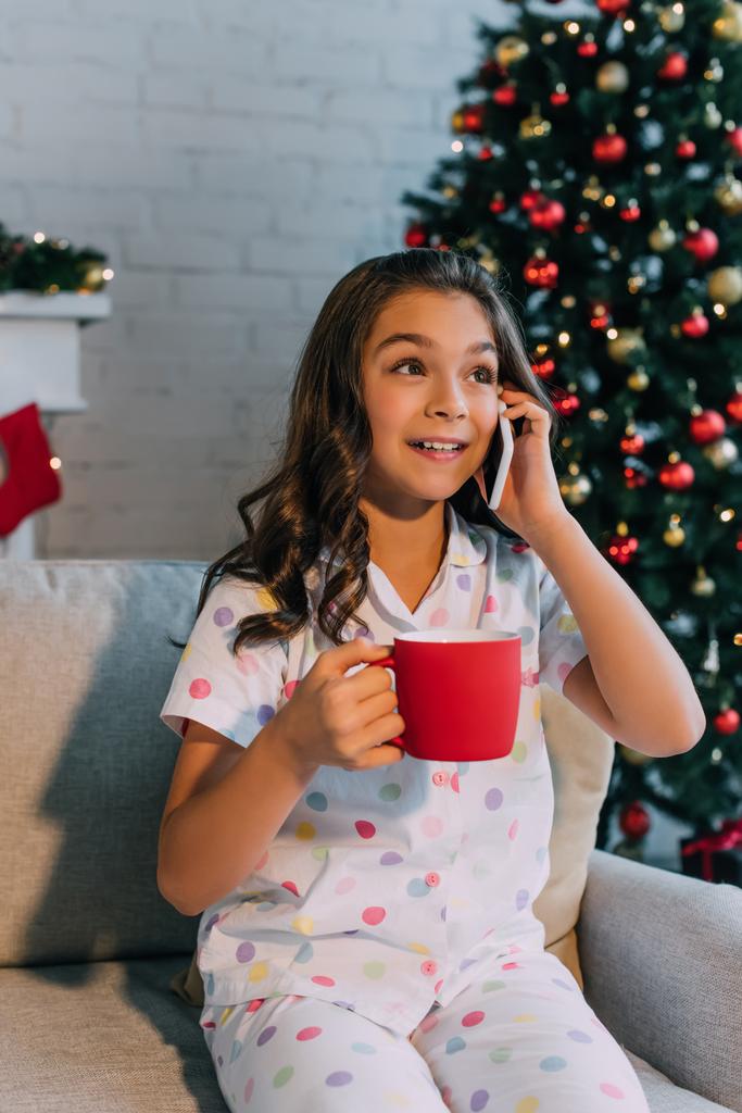 Esiteini lapsi pyjamassa puhuu älypuhelimella ja pitää kuppia joulujuhlan aikana kotona  - Valokuva, kuva