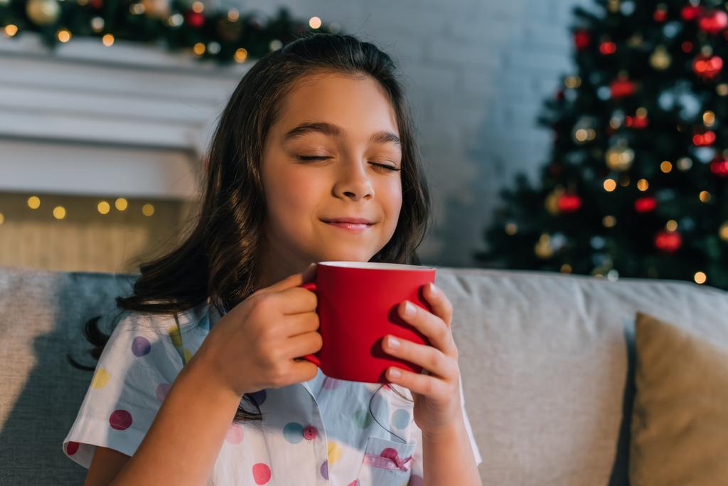 Удовлетворенный ребенок в пижаме нюхает напиток в чашке во время празднования Рождества дома  - Фото, изображение