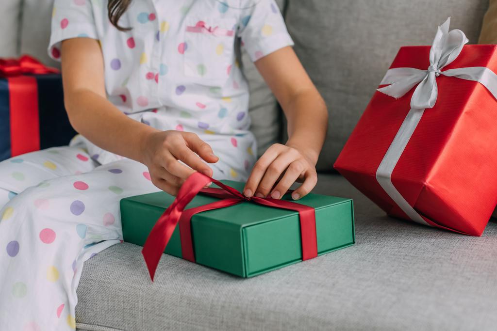Przycięty widok nastolatka w piżamie rozwiązywanie wstążki świąteczny prezent na kanapie  - Zdjęcie, obraz