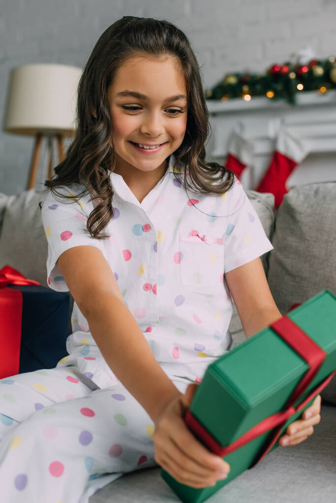 Ragazza felice in pigiama punteggiato tenendo offuscata regalo di Natale sul divano a casa  - Foto, immagini