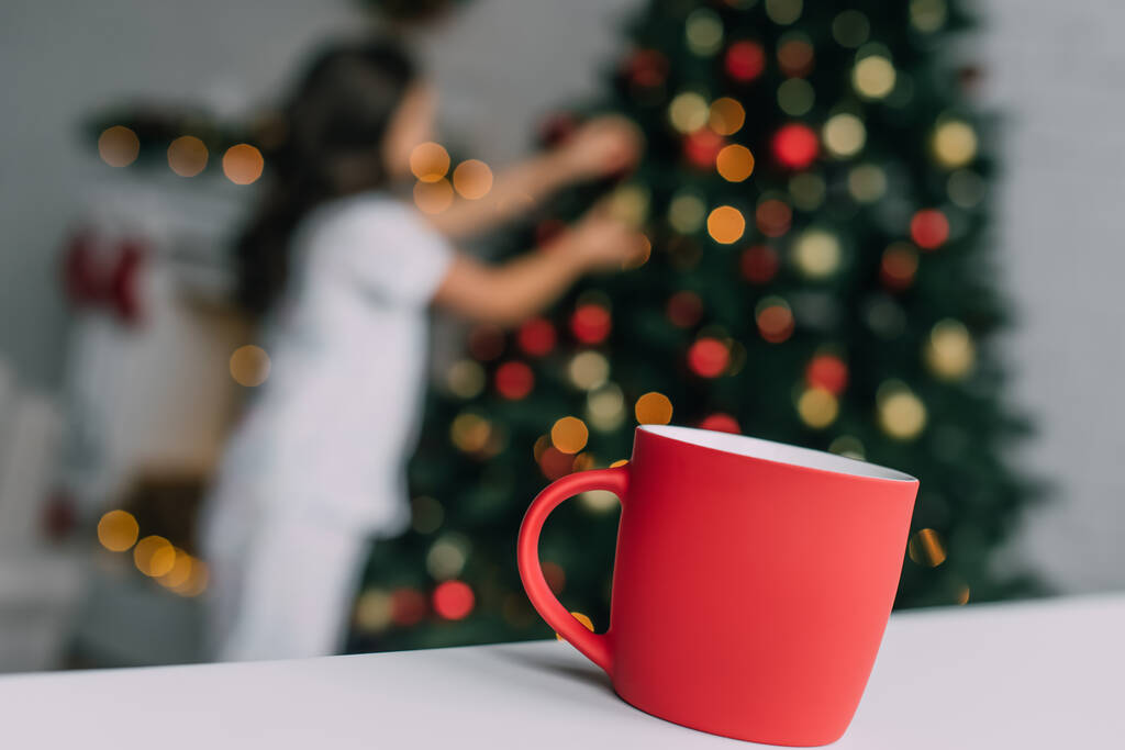 Κόκκινο κύπελλο στο τραπέζι κοντά θολή κορίτσι και χριστουγεννιάτικο δέντρο στο σπίτι  - Φωτογραφία, εικόνα
