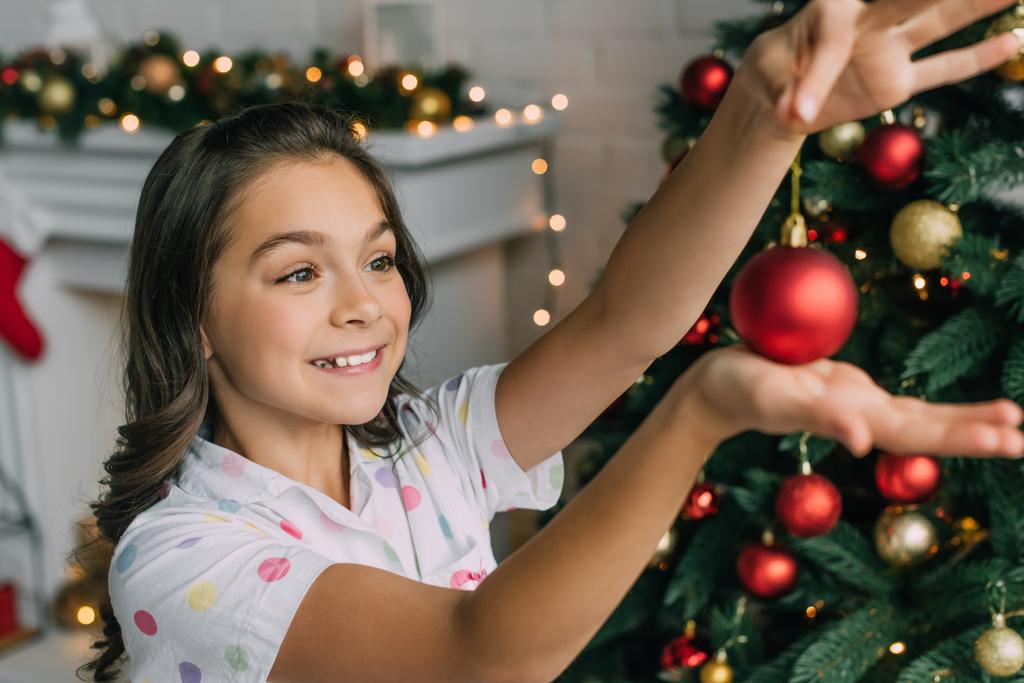 Fille joyeuse en pyjama tenant boule floue près de l'arbre de Noël à la maison  - Photo, image