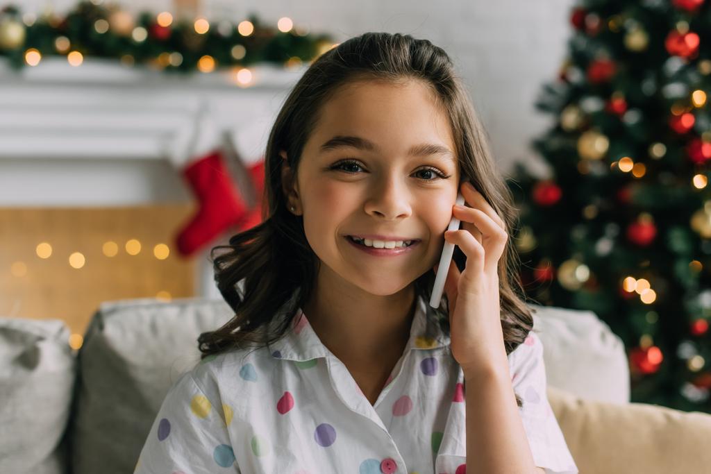 Enfant joyeux en pyjama parlant sur smartphone pendant la célébration à la maison  - Photo, image