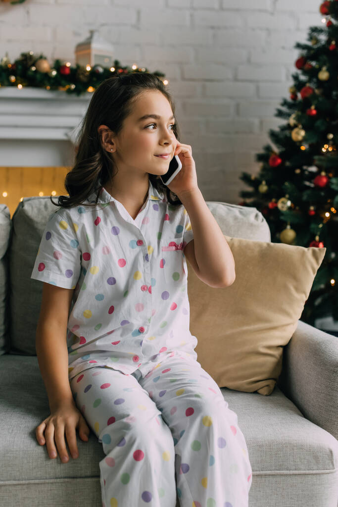 自宅でクリスマスのお祝いの間にソファの上でスマートフォンで話すpajamaの10代の子供  - 写真・画像
