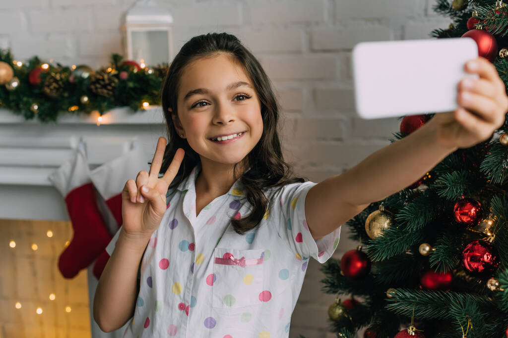 Pijamalı gülümseyen kız Noel ağacının yanındaki akıllı telefondan selfie çekerken barış jesti yapıyor.  - Fotoğraf, Görsel