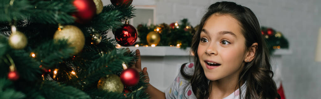 Menina excitada olhando para a árvore de Natal com bugigangas em casa, banner  - Foto, Imagem