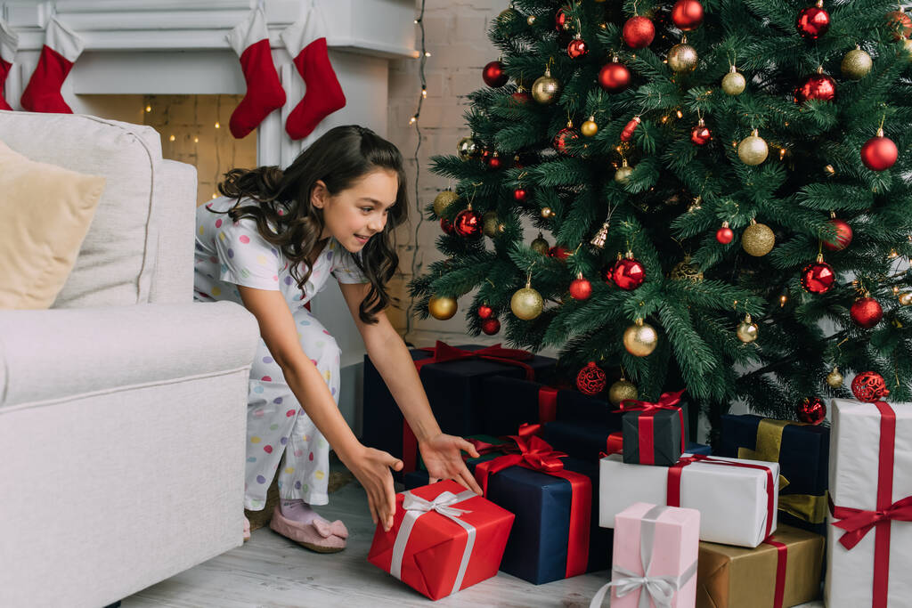 Hymyilevä tyttö pyjamassa ottaa lahja alla joulukuusi olohuoneessa  - Valokuva, kuva