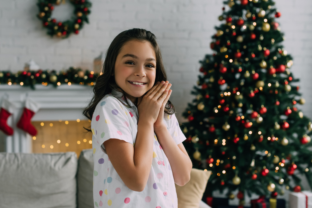 Happy girl in pajama looking at camera and doing praying hands during Christmas celebration at home  - Photo, Image