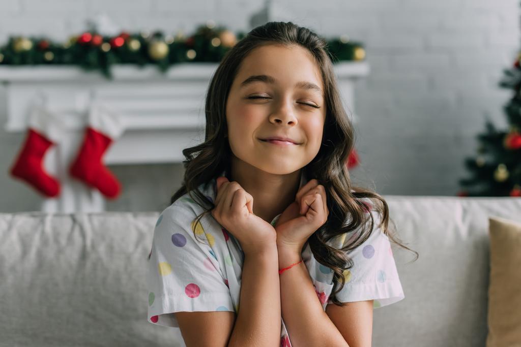Zadowolone dziecko w kropkowanej piżamie siedzi na kanapie podczas świąt Bożego Narodzenia w domu  - Zdjęcie, obraz