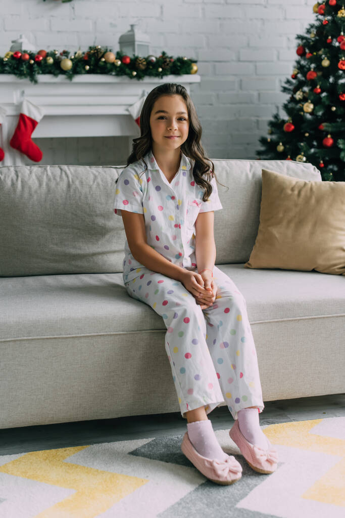 Hymyilevä tyttö katkottu pyjama istuu sohvalla lähellä hämärtynyt takka ja joulukuusi kotona  - Valokuva, kuva