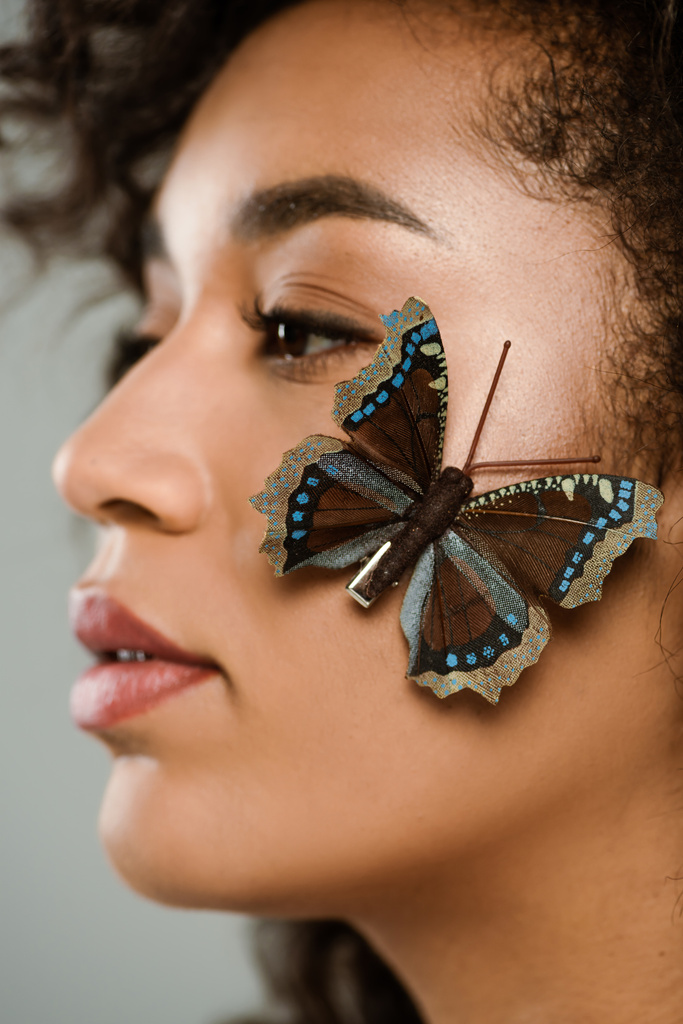 Nahaufnahme von hübschen afrikanisch-amerikanischen Frau mit Schmetterling im Gesicht isoliert auf grau - Foto, Bild