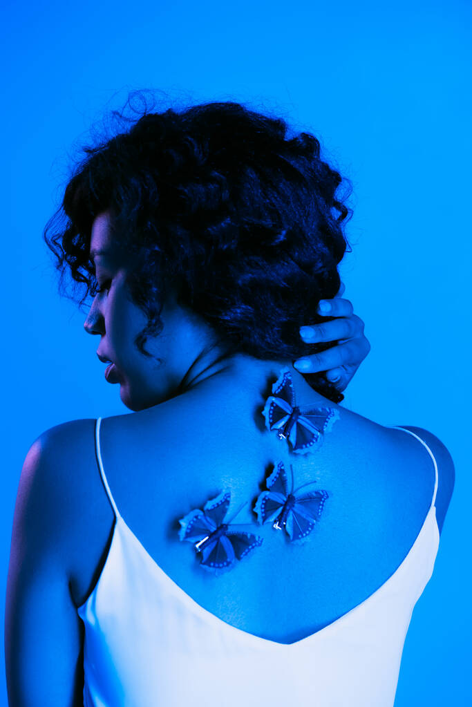 σγουρή αφροαμερικανή γυναίκα με πεταλούδες στην πλάτη ποζάροντας απομονωμένη στο φωτεινό μπλε  - Φωτογραφία, εικόνα