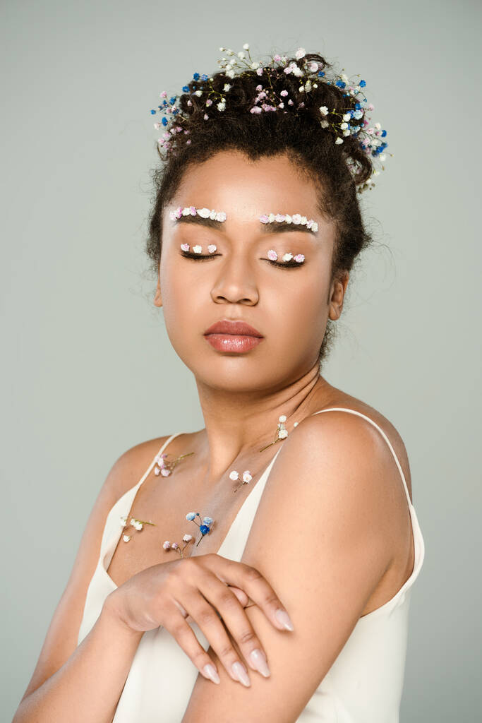 młoda Afroamerykanka z kwiatami na brwiach i zamkniętymi oczami pozuje na szarości - Zdjęcie, obraz