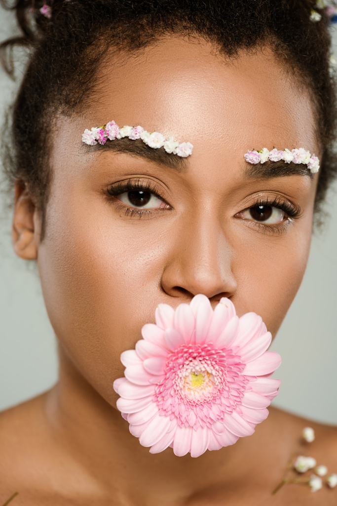 primo piano di donna afroamericana con fiori sulle sopracciglia e capelli con gerbera in bocca isolata sul grigio - Foto, immagini