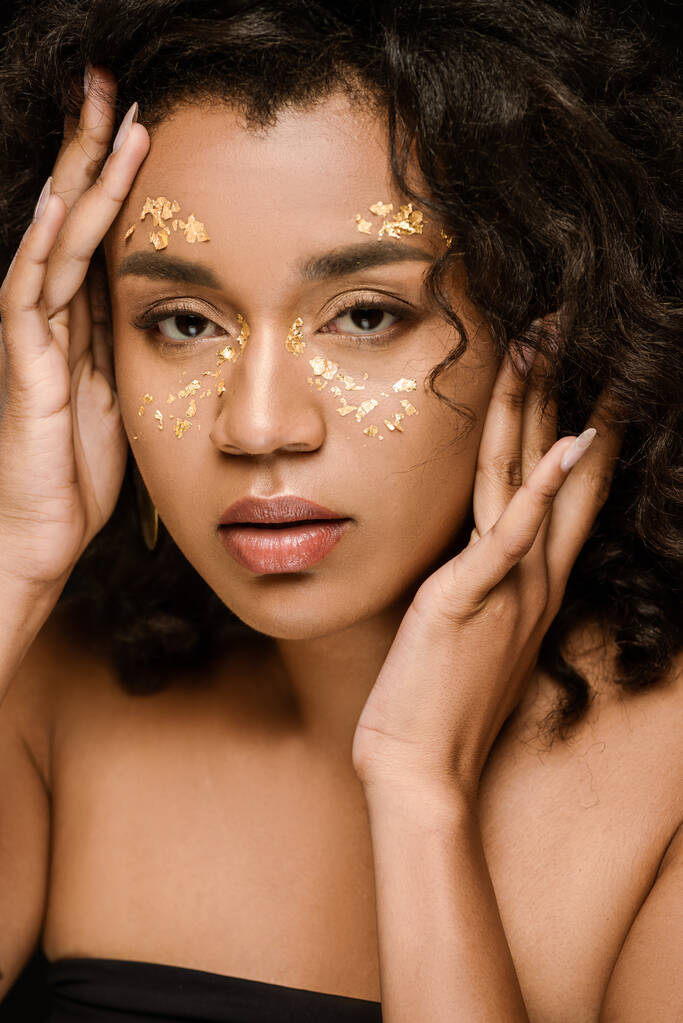 curly african american woman with golden paint on face looking at camera isolated on black - Photo, Image
