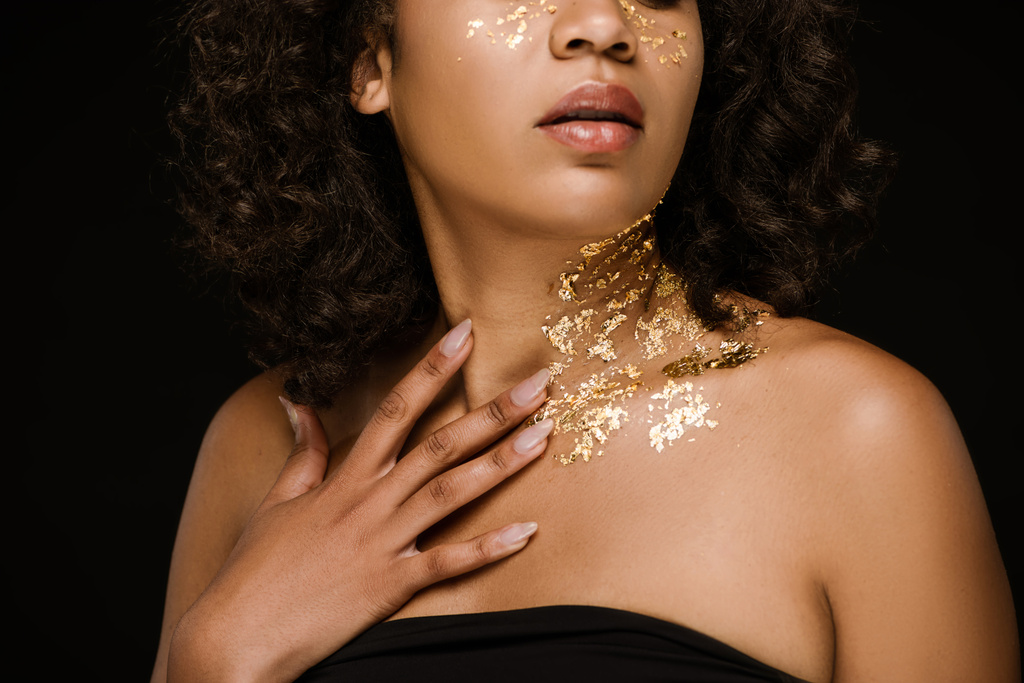 καλλιεργημένη άποψη της νεαρής αφρικανικής αμερικανικής γυναίκας με χρυσή μπογιά στο πρόσωπο και το λαιμό απομονώνονται σε μαύρο - Φωτογραφία, εικόνα