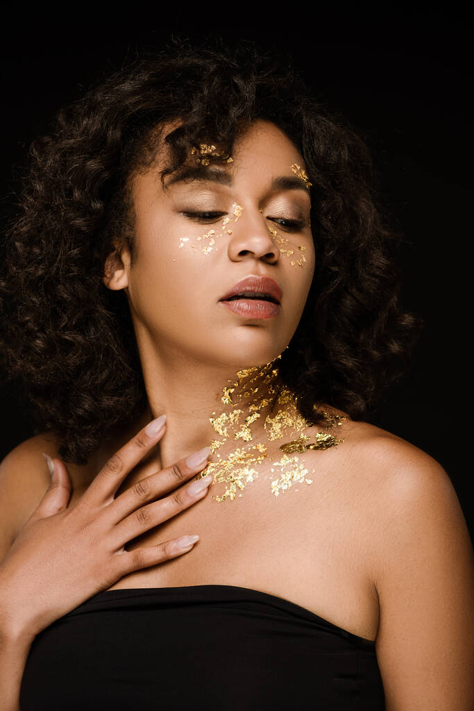 mladá africká americká žena se zlatou barvou na obličeji a krku odvrací pohled izolované na černé - Fotografie, Obrázek