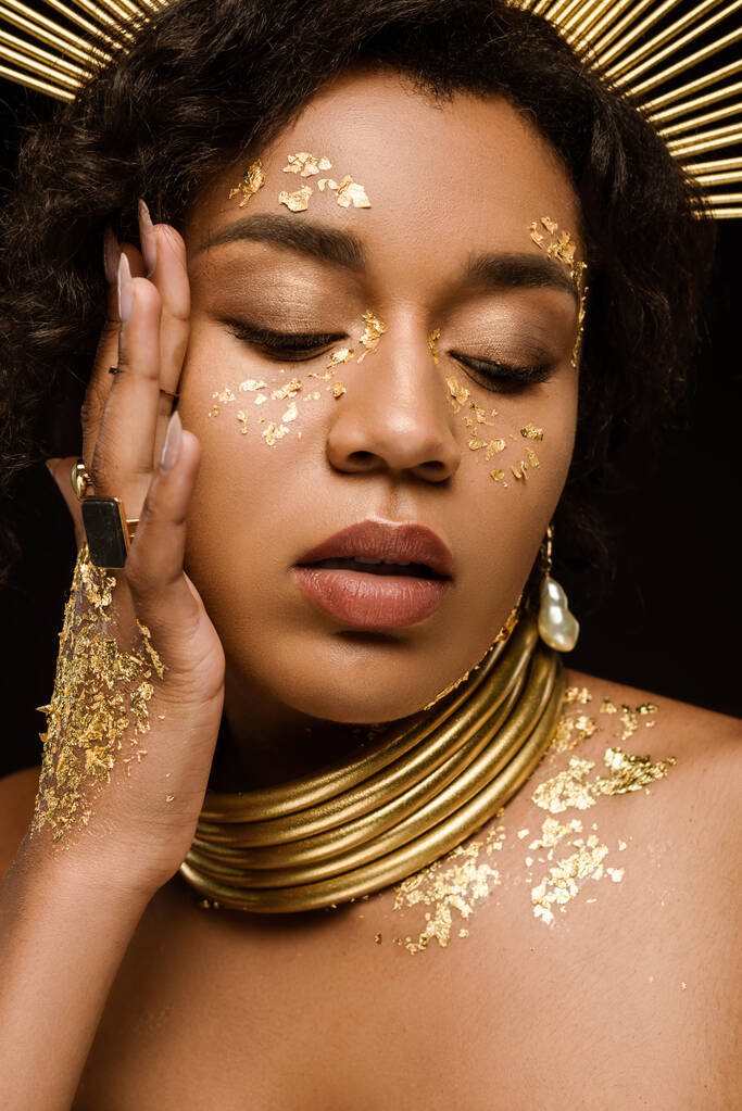 jong Afrikaans Amerikaans model met gouden accessoires en verf op gezicht geïsoleerd op zwart - Foto, afbeelding
