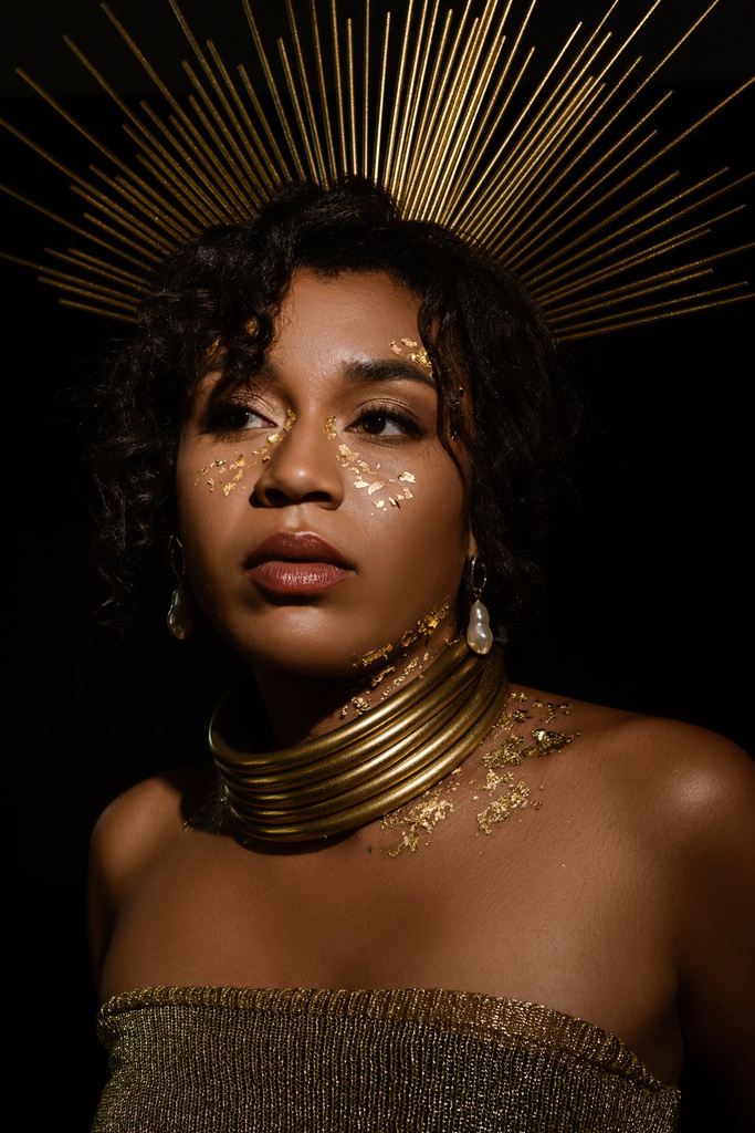 mujer afroamericana confiada con corona dorada y pintura en las mejillas mirando hacia otro lado aislado en negro - Foto, imagen