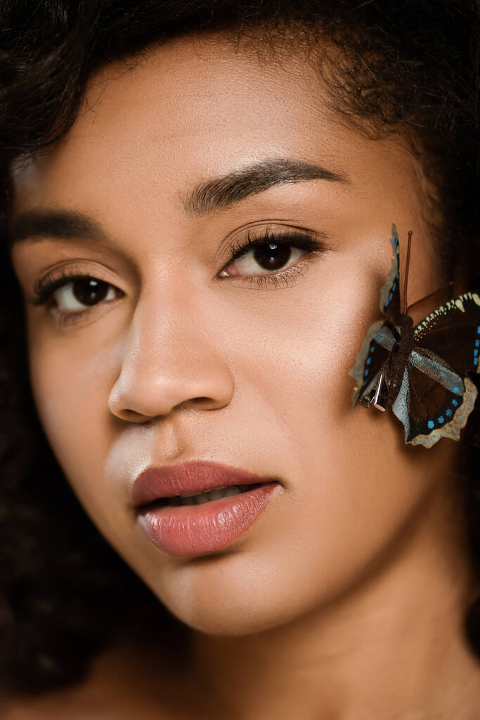 Nahaufnahme einer lockigen Afroamerikanerin mit Schmetterling im Gesicht, die in die Kamera schaut - Foto, Bild