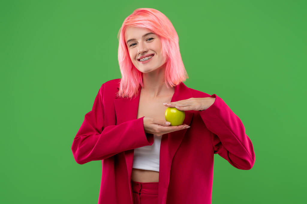 taille-up portret van een vrolijk meisje met een tong piercing en beugels met een groene appel in haar handen - Foto, afbeelding
