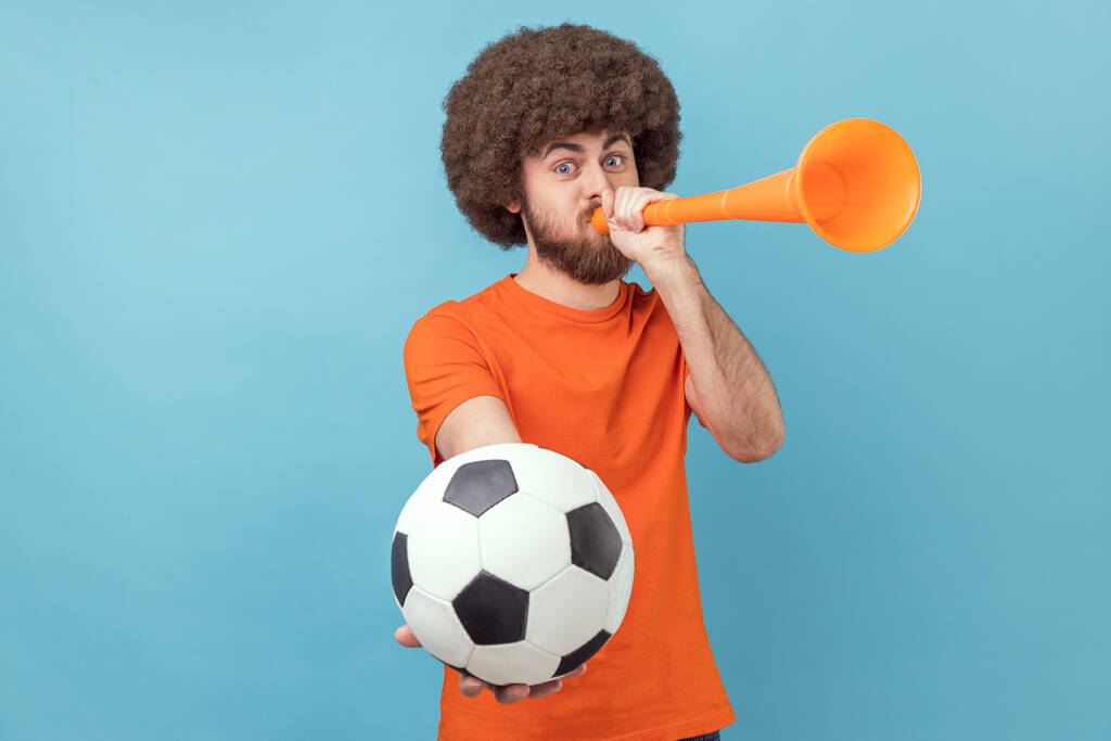 Portrait of man with Afro hairstyle wearing orange T-shirt celebrating victory of favourite football team, blowing horn and ball in hands. Indoor studio shot isolated on blue background. - Photo, Image