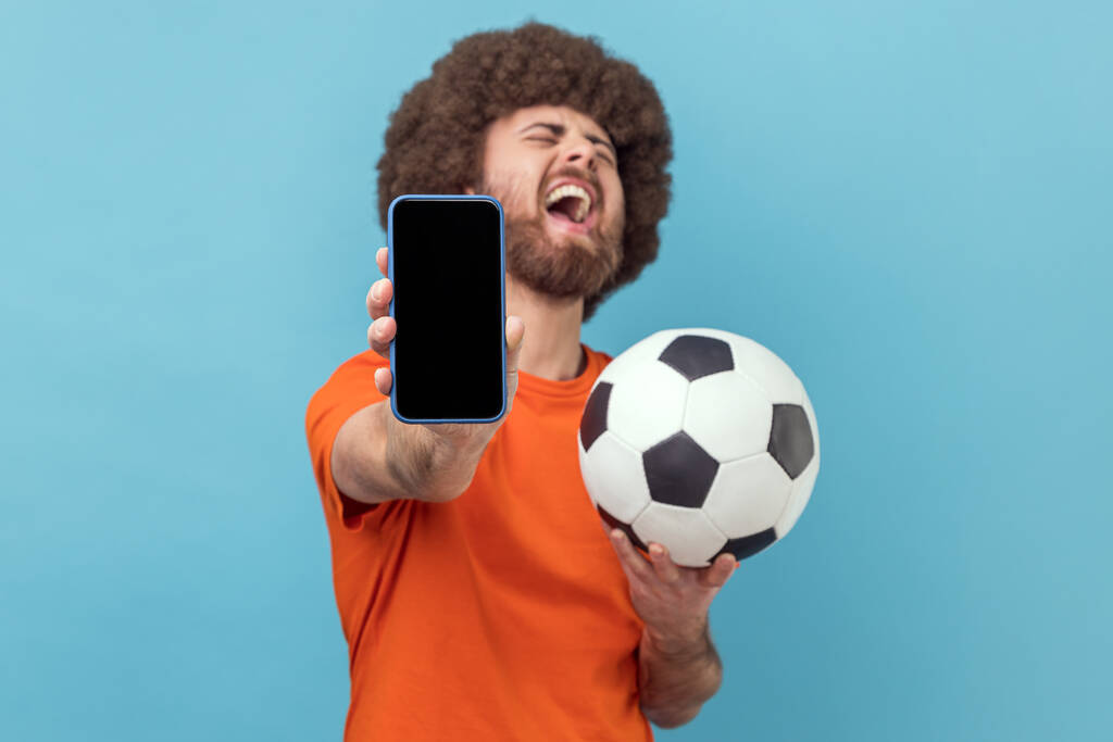 Son derece mutlu, Afro saçlı, tişörtlü futbol topuyla ayakta duran, cep telefonunu boş gösteren, bahis oynayan ve kazanan bir adam. Mavi arka planda kapalı stüdyo çekimi. - Fotoğraf, Görsel